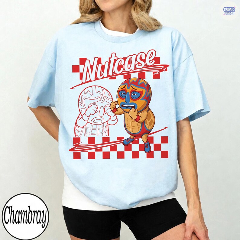 May include: A light blue t-shirt featuring a graphic of a peanut character wearing a wrestling mask and outfit. The text "Nutcase" appears above the character, set against a red and white checkered background. The design includes a line drawing of a second peanut character.
