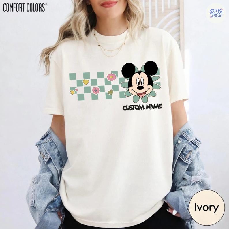 May include: Ivory-colored t-shirt with a Mickey Mouse graphic. The design includes a checkerboard pattern with flowers and hearts. The text "CUSTOM NAME" is below the character. The shirt is from Comfort Colors.
