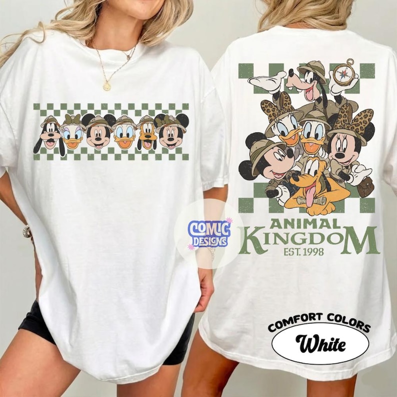 Pode incluir: Camiseta branca com um gr&aacute;fico de personagens da Disney em trajes de safari. A frente exibe uma fileira de personagens acima de um padr&atilde;o xadrez verde e branco. A parte de tr&aacute;s tem um grupo maior de personagens com o texto "Animal Kingdom".