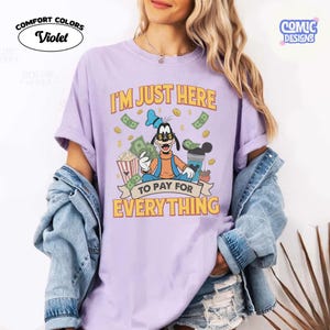 May include: Violet Comfort Colors t-shirt featuring a cartoon Goofy character holding money, popcorn, and a drink. The shirt has the text "I'M JUST HERE TO PAY FOR EVERYTHING" in yellow and orange. The shirt is a light purple color.