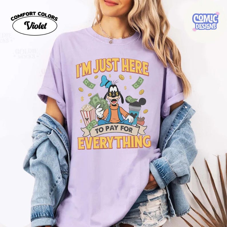 May include: Violet Comfort Colors t-shirt featuring a cartoon Goofy character holding money, popcorn, and a drink. The shirt has the text "I'M JUST HERE TO PAY FOR EVERYTHING" in yellow and orange. The shirt is a light purple color.