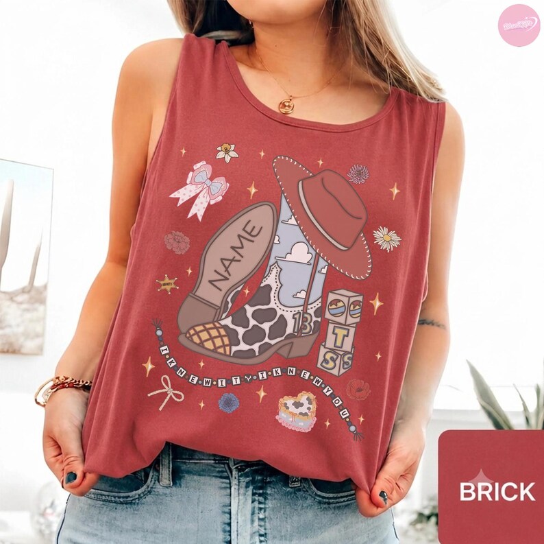 Puede incluir: Una camiseta sin mangas de color rojo ladrillo con un estampado gr&aacute;fico de una bota de vaquero con patr&oacute;n de manchas de vaca, un sombrero marr&oacute;n y bloques. La bota incluye el texto NAME y el n&uacute;mero 13. Otros gr&aacute;ficos incluyen lazos, flores y estrellas.