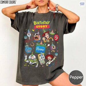 May include: A charcoal gray short-sleeve t-shirt featuring a colorful graphic print with the text BIRTHDAY STORY and Name. The design includes various cartoon characters wearing party hats, a red balloon with the number 5, and confetti.
