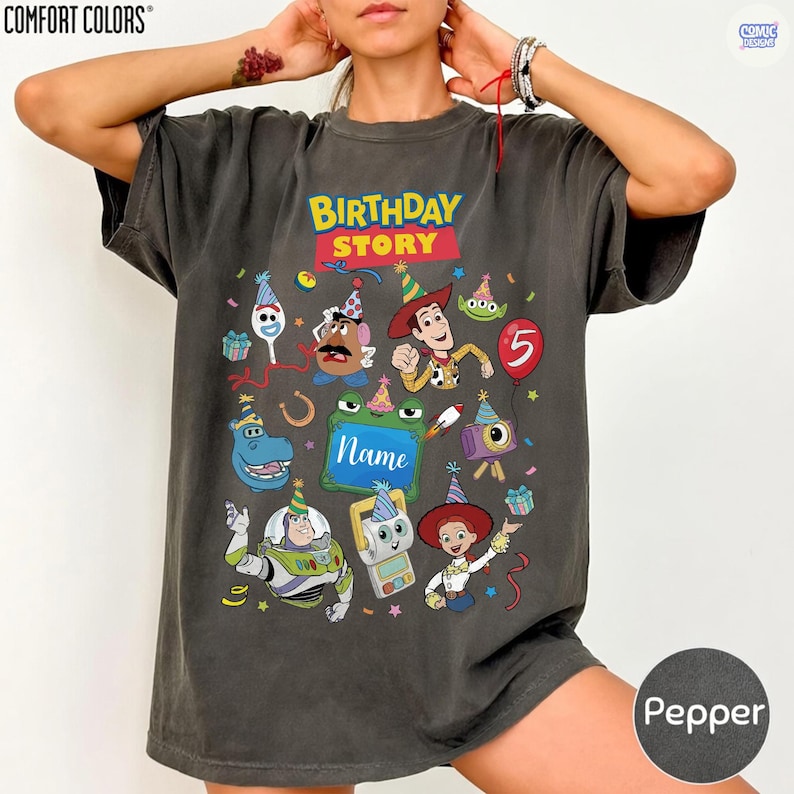May include: A charcoal gray short-sleeve t-shirt featuring a colorful graphic print with the text BIRTHDAY STORY and Name. The design includes various cartoon characters wearing party hats, a red balloon with the number 5, and confetti.
