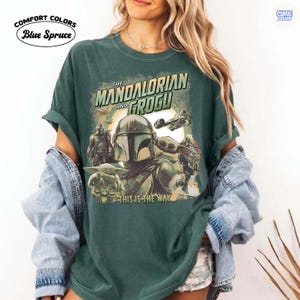 May include: A dark green t-shirt featuring a graphic of The Mandalorian and Grogu, with the text "THE MANDALORIAN AND GROGU" and "THIS IS THE WAY". The shirt has rolled-up sleeves and the words "Comfort Colors Blue Spruce" are printed on the upper left.