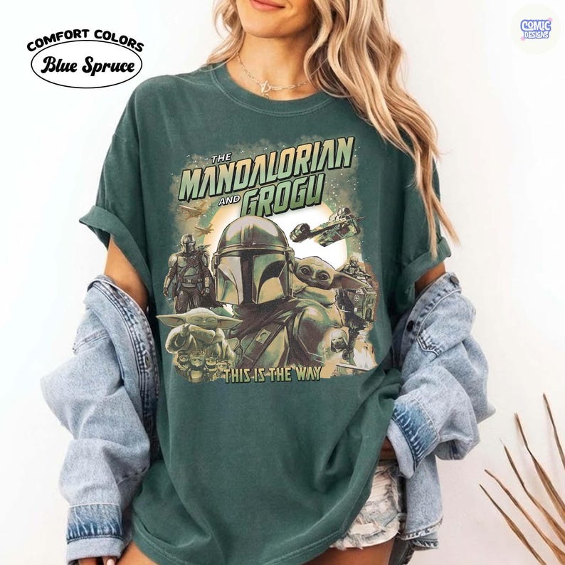 May include: A dark green t-shirt featuring a graphic of The Mandalorian and Grogu, with the text "THE MANDALORIAN AND GROGU" and "THIS IS THE WAY". The shirt has rolled-up sleeves and the words "Comfort Colors Blue Spruce" are printed on the upper left.