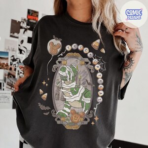 Puede incluir: Una camiseta negra con un gr&aacute;fico de un dinosaurio verde envuelto en vendas blancas como una momia. El dise&ntilde;o incluye el texto "TRICK OR TREAT", un castillo, murci&eacute;lagos, dulces de ma&iacute;z y dos cabezas de rat&oacute;n con forma de calabaza con 19 y 95.