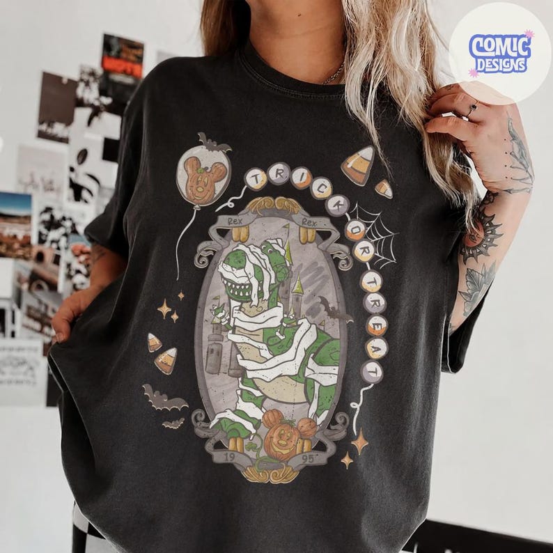 Puede incluir: Una camiseta negra con un gr&aacute;fico de un dinosaurio verde envuelto en vendas blancas como una momia. El dise&ntilde;o incluye el texto "TRICK OR TREAT", un castillo, murci&eacute;lagos, dulces de ma&iacute;z y dos cabezas de rat&oacute;n con forma de calabaza con 19 y 95.