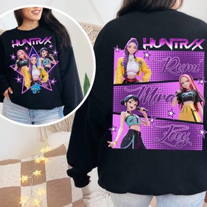 Peut inclure: Sweat-shirt noir avec le texte "HUNTRAX" et des illustrations de personnages animés. Le devant présente un motif en forme d'étoile. Le dos affiche les noms "Rumi", "Mina" et "Joey" avec des images de personnages.