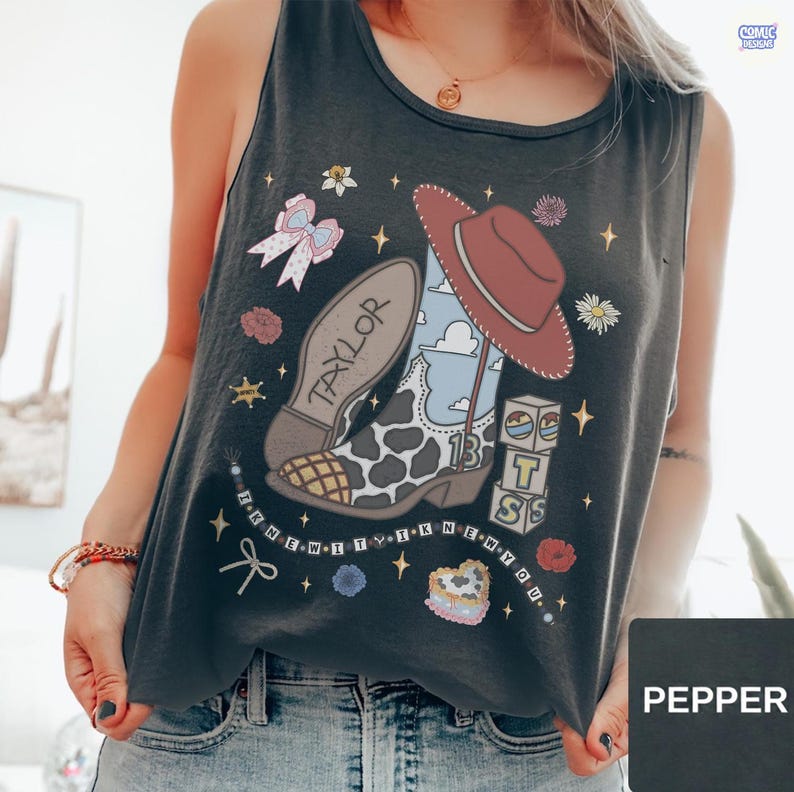 Puede incluir: Una camiseta sin mangas de color gris carb&oacute;n con un estampado gr&aacute;fico de una bota de vaquero con manchas de vaca, un sombrero de vaquero rojo, un lazo rosa y bloques de letras que forman T-S. El dise&ntilde;o incluye estrellas y flores.