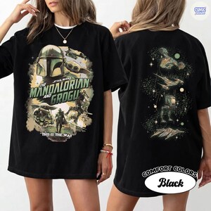 Op de afbeelding: Zwart T-shirt met een ontwerp van The Mandalorian en Grogu. De voorkant heeft een afbeelding met de tekst "THIS IS THE WAY". De achterkant toont schepen en personages in een ruimtethema.