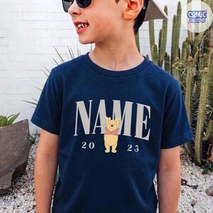 K&ouml;nnte beinhalten: Marineblaues T-Shirt mit dem Wort "NAME" in hellgrauer, fetter Schrift. Darunter befindet sich ein Cartoon-Bild von Winnie the Pooh, mit den Zahlen "20" und "23" auf beiden Seiten. Das T-Shirt hat einen Rundhalsausschnitt.