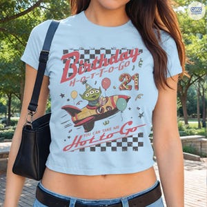 May include: Light blue crop top with a graphic featuring an alien in a rocket ship, balloons, and the text "Birthday H-O-T-T-O-GO 21". The shirt also includes the text "You can take me Hot to Go".