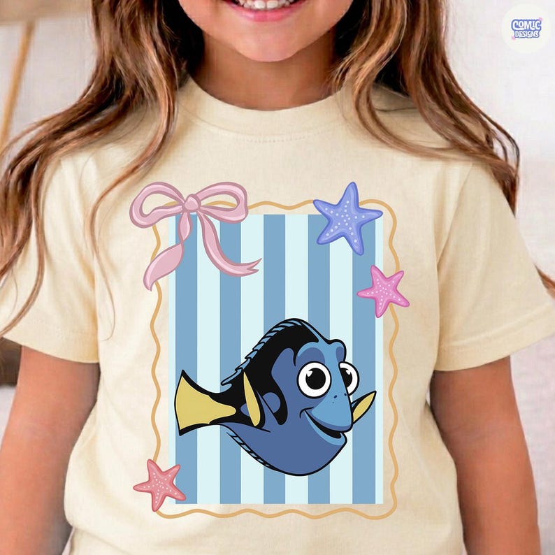 May include: A light yellow t-shirt featuring a cartoon image of a blue fish with yellow fins, set against a blue and white striped background. The design includes a pink bow and starfish. The text "COMIC DESIGNS" is in the upper right corner.