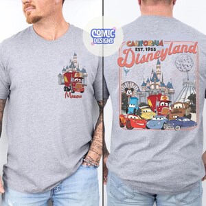 K&ouml;nnte beinhalten: Graues T-Shirt mit einem Cartoon-Design eines roten Trucks und Schlosses auf der Vorderseite, mit dem Namen "Mason". Die R&uuml;ckseite zeigt ein Disneyland-Design mit dem Text "California Est. 1955 Disneyland" und Cartoon-Autos.