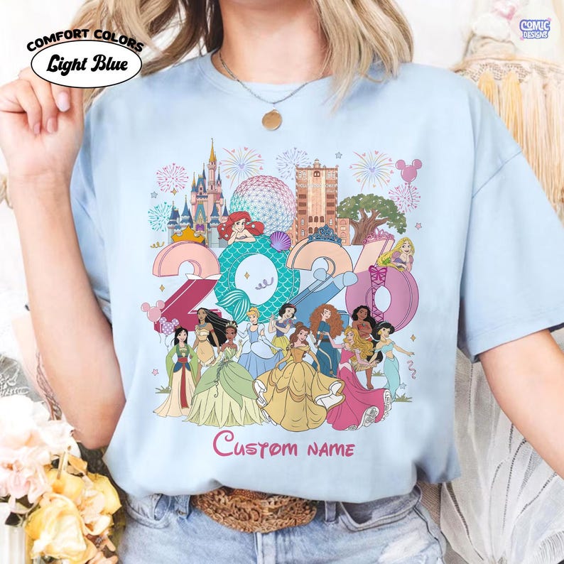 May include: A light blue t-shirt featuring a graphic print of Disney princesses, a castle, and the year 2024. The design includes fireworks, park icons, and the text Custom name in pink script. The shirt is labeled as Comfort Colors Light Blue.