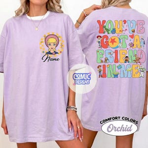 Op de afbeelding: Een orchideekleurig t-shirt met een afbeelding op de voorkant van een blond personage in een cirkel met de tekst Name. De achterkant toont een kleurrijke collage van diverse geanimeerde personages en de tekst YOU'VE GOT A FRIEND IN ME.