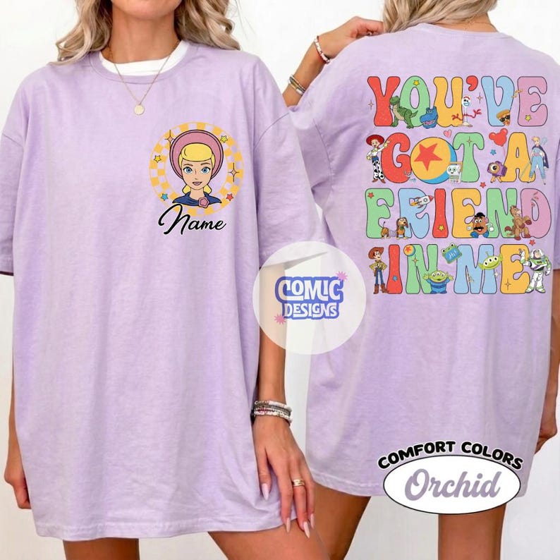 Op de afbeelding: Een orchideekleurig t-shirt met een afbeelding op de voorkant van een blond personage in een cirkel met de tekst Name. De achterkant toont een kleurrijke collage van diverse geanimeerde personages en de tekst YOU'VE GOT A FRIEND IN ME.
