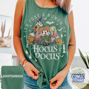May include: A light green sleeveless tank top featuring a graphic print of three cartoon characters dressed in costumes with pumpkins, bats, and stars. The text reads IT'S JUST A BUNCH OF HOCUS POCUS in white lettering across the front of the garment.