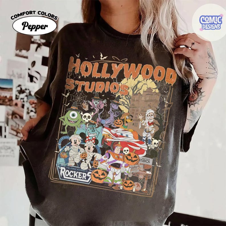 K&ouml;nnte beinhalten: Ein anthrazitfarbenes T-Shirt mit einem Grafikdruck, der den Text HOLLYWOOD STUDIOS und ROCKERS zeigt. Das Design umfasst verschiedene Zeichentrickfiguren in Halloween-Kost&uuml;men, K&uuml;rbisse, Spinnennetze und die Silhouette eines Spukhauses.