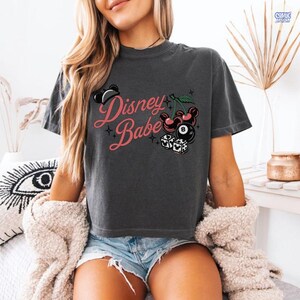 May include: Dark grey t-shirt featuring the text "Disney Babe" in pink script, with Mickey Mouse imagery, cherries, an 8-ball, and dice. The top is a casual, short-sleeved design.