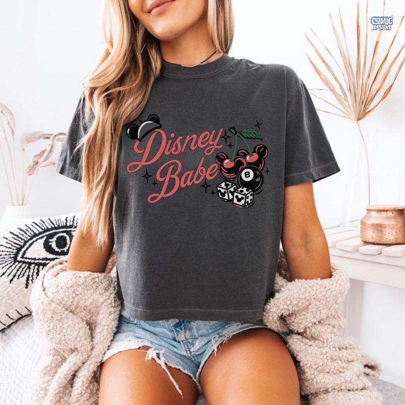 May include: Dark grey t-shirt featuring the text "Disney Babe" in pink script, with Mickey Mouse imagery, cherries, an 8-ball, and dice. The top is a casual, short-sleeved design.