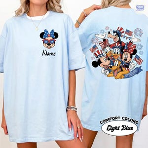 May include: Light blue t-shirt featuring a patriotic Minnie Mouse design with sunglasses and a bow. The back of the shirt displays a group of Disney characters celebrating with flags and fireworks. The text "Name" is on the front.