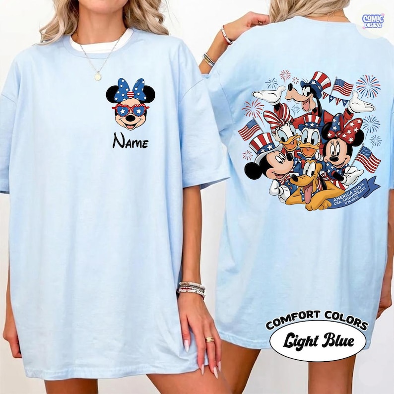 May include: Light blue t-shirt featuring a patriotic Minnie Mouse design with sunglasses and a bow. The back of the shirt displays a group of Disney characters celebrating with flags and fireworks. The text "Name" is on the front.