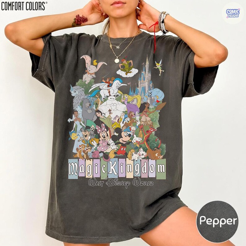 Puede incluir: Camiseta color pimienta con un gr&aacute;fico colorido de personajes de Disney, incluyendo Mickey Mouse, Minnie Mouse y varias princesas. El texto "Magic Kingdom Walt Disney World" est&aacute; impreso debajo. La camiseta es de la marca Comfort Colors.