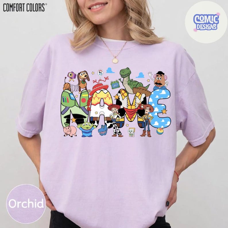 May include: A light purple t-shirt featuring a large graphic print of the word NAME, with each letter stylised to incorporate various Toy Story characters including Woody, Jessie, Buzz Lightyear, Rex, Slinky Dog, and Mr. Potato Head.