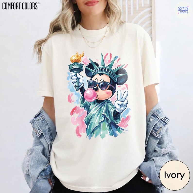 May include: An ivory short-sleeve t-shirt featuring a graphic of Mickey Mouse dressed as the Statue of Liberty. The character wears sunglasses, blows a pink bubble, holds a torch, and makes a peace sign, set against colorful abstract paint splatters.