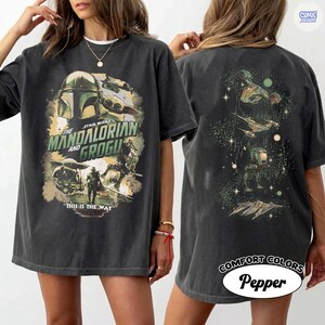 Op de afbeelding: Donkergrijs t-shirt met een afbeelding van "The Mandalorian and Grogu". De voorkant toont de Mandalorian helm en Grogu, met de tekst "THIS IS THE WAY". De achterkant toont ruimteschepen en personages. Het shirt is gelabeld "COMFORT COLORS Pepper."