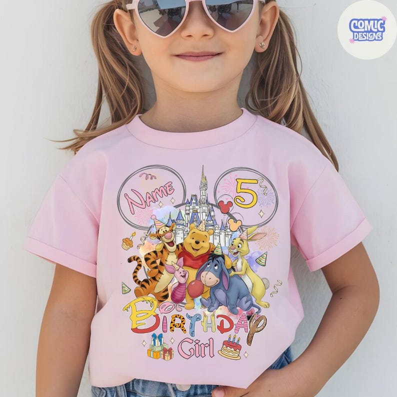 以下が含まれることがあります： ピンク色のTシャツ。くまのプーさんと仲間たちのカラフルなグラフィックが特徴で、「Birthday Girl」の文字と数字の「5」が描かれています。デザインにはお城、風船、ケーキが含まれています。Tシャツは半袖です。