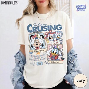 以下が含まれることがあります： アイボリー色のTシャツ。ミニーマウスとドナルドダックがセーラー服を着たグラフィックで、「Girl goes Cruising」の文字が入っています。デザインには額縁に入った画像と航海要素が含まれています。このシャツはComfort Colors製です。