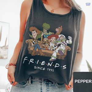 May include: A dark gray sleeveless tank top featuring a graphic print of various animated characters sitting on a couch. Below the illustration, the white text reads "F R I E N D S" with colorful dots between the letters, followed by "SINCE 1995".