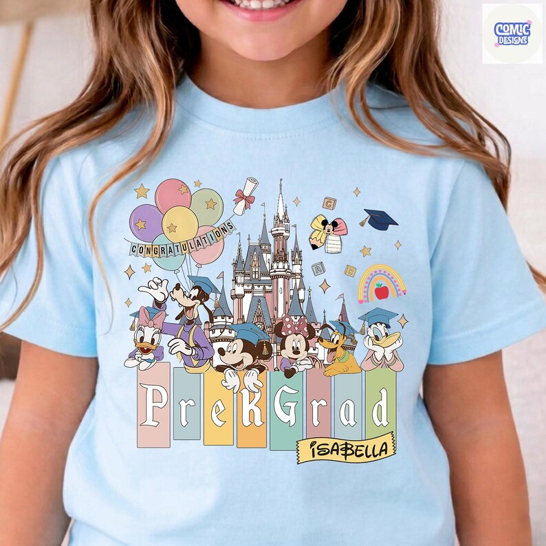 May include: Light blue t-shirt featuring a colorful graphic with Disney characters, a castle, balloons, and the text "Congratulations." Below, the shirt reads "Prek Grad" with the name "Isabella." The design includes graduation caps and a rainbow.
