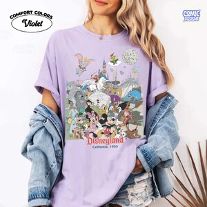 Puede incluir: Camiseta Violet Comfort Colors con un dise&ntilde;o de Disneyland. El dise&ntilde;o presenta a Mickey y Minnie Mouse, Dumbo y otros personajes cl&aacute;sicos de Disney. Tambi&eacute;n se puede ver el texto "Disneyland California, 1955".