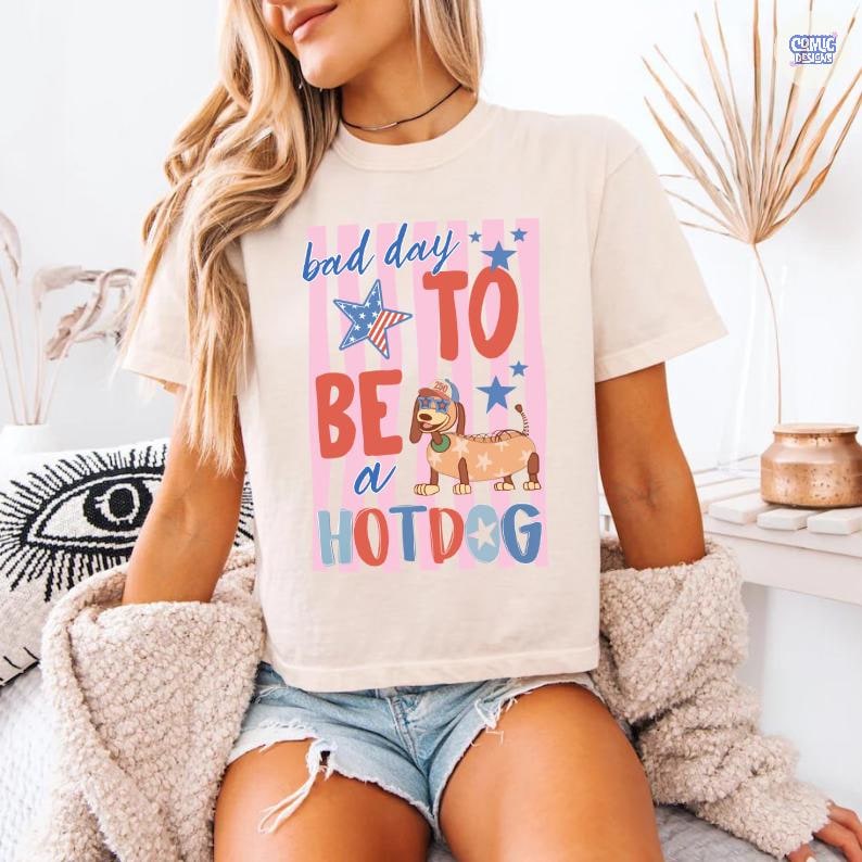 May include: Cream-colored t-shirt with a graphic that reads "bad day to be a hotdog." The graphic features a dachshund wearing sunglasses and a hotdog costume, with stars and stripes in red, white, and blue.