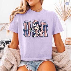 Op de afbeelding: Een lichtpaars t-shirt met een afbeelding van Mickey Mouse met een patriottische hoge hoed. De tekst luidt 250TH ANNIVERSARY USA 1776 2026. De letters USA zijn gevuld met rode en witte strepen en blauwe sterren, geaccentueerd door blauwe strikken.
