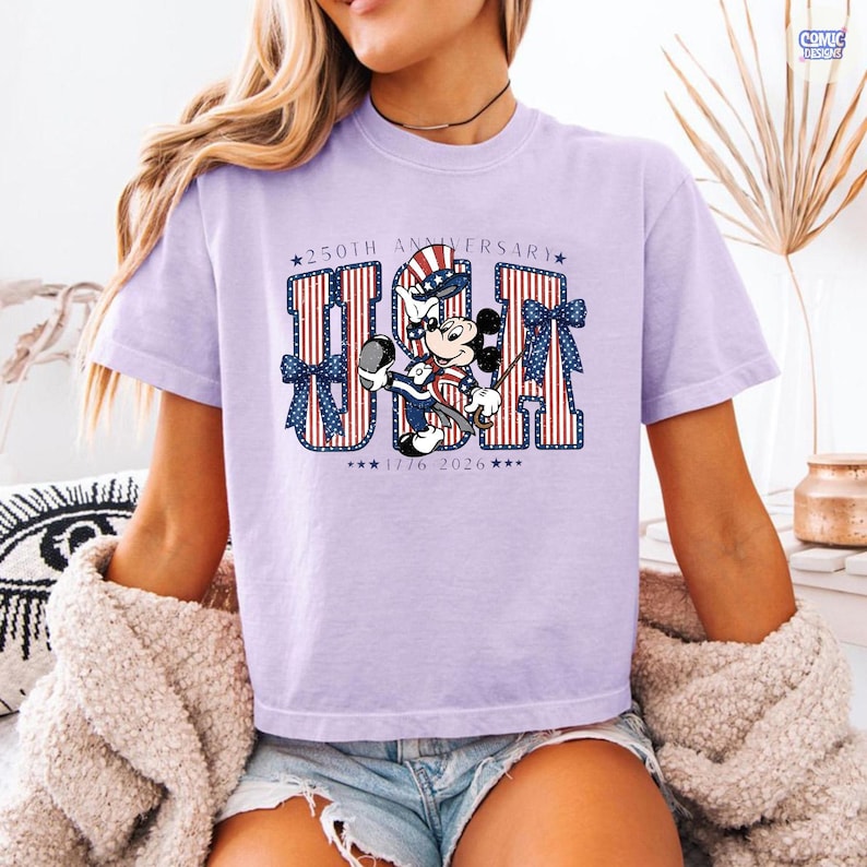 Op de afbeelding: Een lichtpaars t-shirt met een afbeelding van Mickey Mouse met een patriottische hoge hoed. De tekst luidt 250TH ANNIVERSARY USA 1776 2026. De letters USA zijn gevuld met rode en witte strepen en blauwe sterren, geaccentueerd door blauwe strikken.