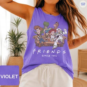 May include: A purple sleeveless tank top featuring a graphic print of Toy Story characters sitting on a couch with the text "FRIENDS SINCE 1995" printed below the illustration. The characters include Woody, Rex, Bullseye, Jessie, and Buzz Lightyear.