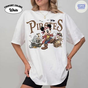 May include: White Comfort Colors t-shirt featuring a cartoon Mickey Mouse pirate design. The design includes the word "Pirates" in brown lettering, a pirate ship, treasure chest, and Mickey Mouse in a pirate outfit. The shirt also has the words "Comfort Colors" and "White".
