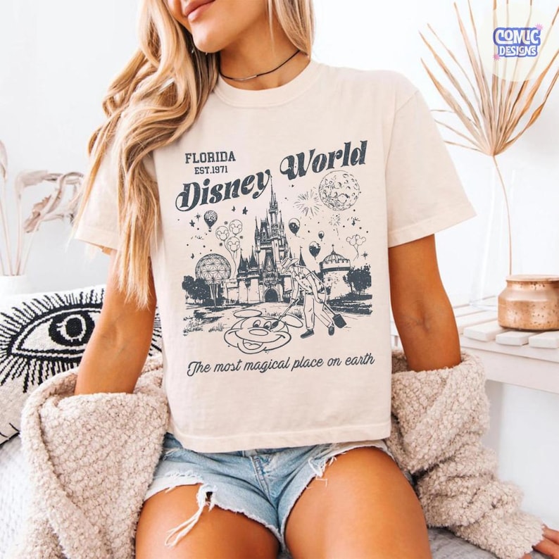 K&ouml;nnte beinhalten: Cremefarbenes T-Shirt mit einer Grafik von Disney World in Florida. Das Design zeigt ein Schloss, Mickey Mouse und den Text "Florida Est. 1971 Disney World" und "The most magical place on earth."