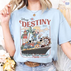 Puede incluir: Camiseta azul claro con un dise&ntilde;o gr&aacute;fico de personajes de Disney en un crucero. El dise&ntilde;o incluye un faro, palmeras y el texto "Disney DESTINY 2026".