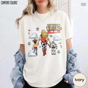 May include: An ivory short-sleeve t-shirt featuring a graphic of three cartoon characters walking away, with the text "YOU'VE GOT A FRIEND IN ME". The design includes a cowboy, a cowgirl with a guitar, and a space ranger, surrounded by clouds and stars.