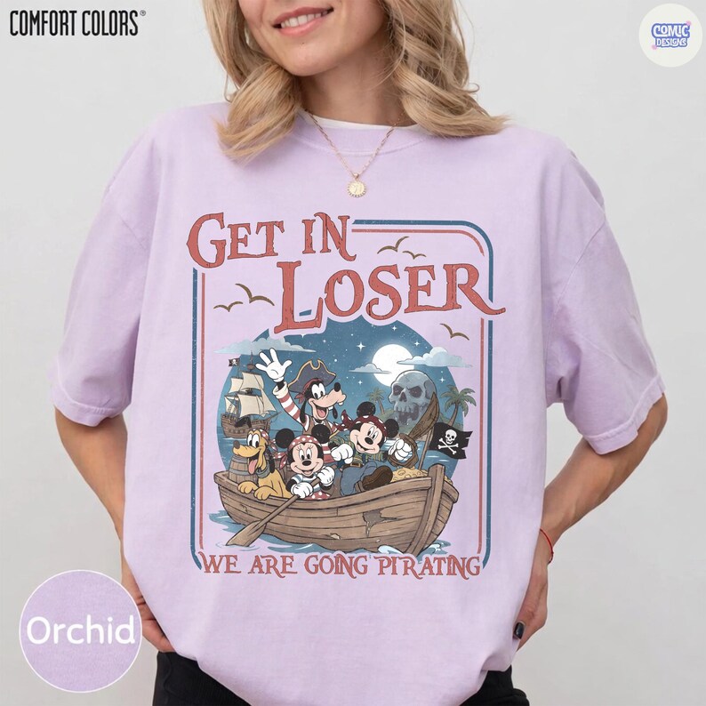 May include: Orchid-colored t-shirt with a pirate-themed graphic. The design features cartoon characters in a boat with the text "Get In Loser" and "We Are Going Pirating". The shirt is a Comfort Colors brand.