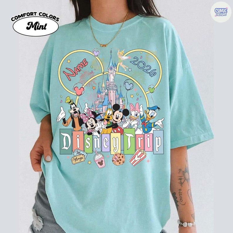 May include: Mint green Comfort Colors t-shirt featuring a Disney Trip design. The design includes Disney characters, a castle, and the text "Name" and "2020". Additional graphics include fireworks, hearts, and stars.