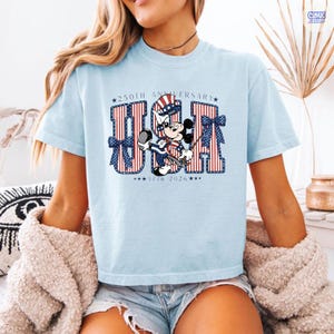 Op de afbeelding: Een lichtblauw t-shirt met een afbeelding van Mickey Mouse die een patriottische hoge hoed draagt. Het ontwerp bevat de tekst 250TH ANNIVERSARY USA 1776 2026, met grote rood-wit gestreepte letters versierd met blauwe strikken en witte sterren.