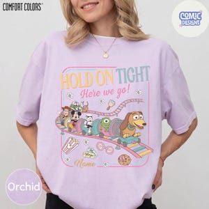 May include: Orchid-colored t-shirt with a colorful graphic of a roller coaster featuring cartoon characters. The text "HOLD ON TIGHT Here we go!" is displayed above the roller coaster. The shirt has the "COMFORT COLORS" and "COMIC DESIGNS" logos.