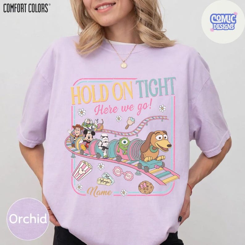 May include: Orchid-colored t-shirt with a colorful graphic of a roller coaster featuring cartoon characters. The text "HOLD ON TIGHT Here we go!" is displayed above the roller coaster. The shirt has the "COMFORT COLORS" and "COMIC DESIGNS" logos.